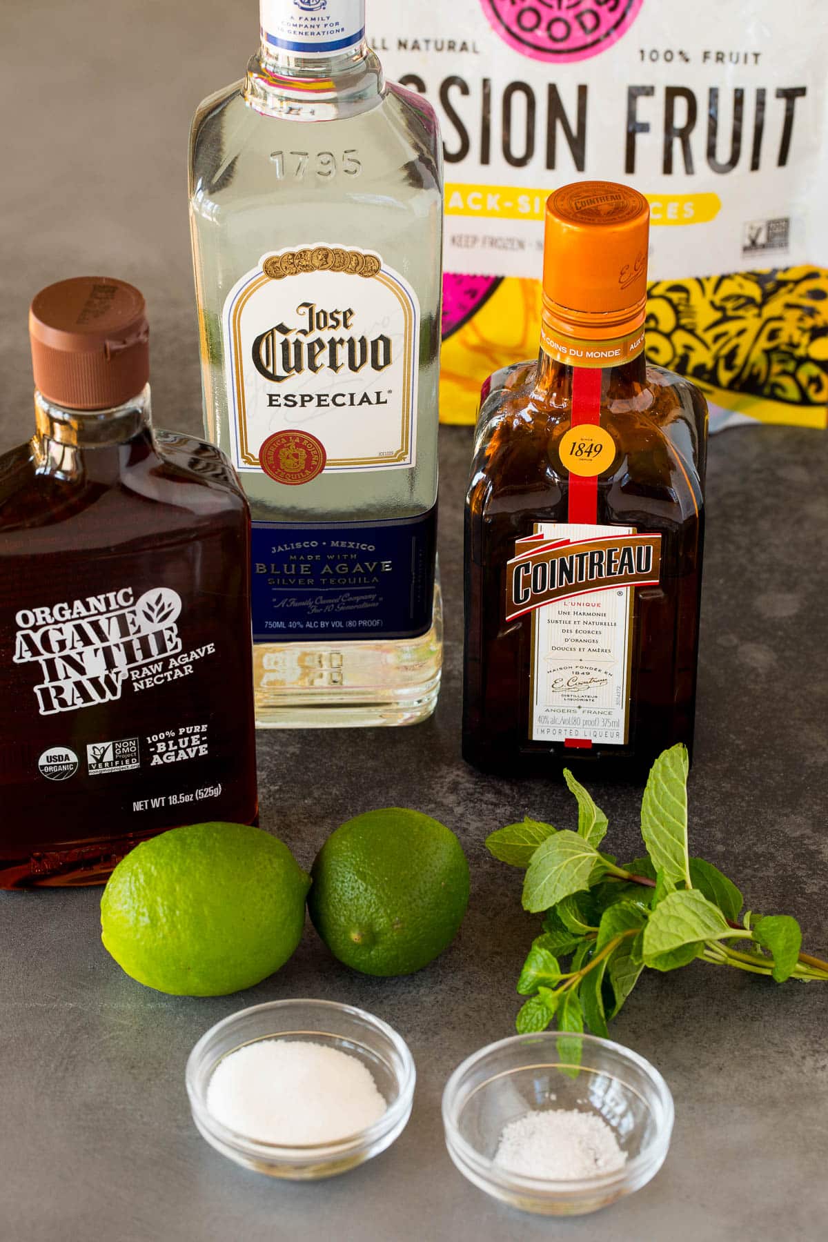 Ingredients including bottles of tequila, triple sec, passion fruit puree, agave, limes and mint.