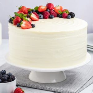 A berry chantilly cake on a cake stand, topped with fresh berries and mint sprigs.
