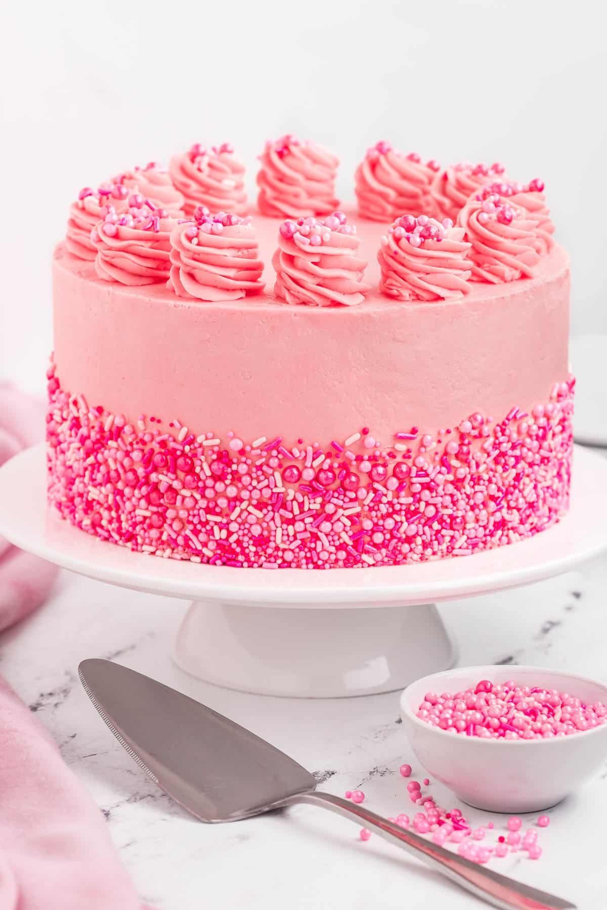 A pink velvet cake on a pedestal decorated with sprinkles.