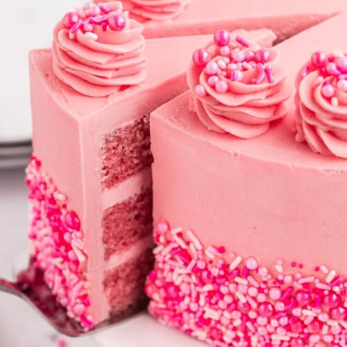 A pink cake with a slice being served out of it. The pink cake is decorated with sprinkles.