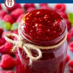 A jar of raspberry sauce surrounded by fresh raspberries.