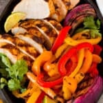 A platter of grilled chicken fajitas with onions, bell peppers, lime wedges and fresh cilantro.