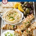 Chicken souvlaki skewers on a plate with marinated olives, hummus ant tzatziki.