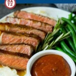 A bowl of steak sauce on a plate with steak, mashed potatoes and green beans.