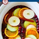 A slow cooker full of apple cider garnished with apple slices, orange slices, spices and cranberries.